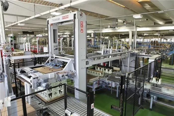 An OCME in-line palletizer with a layer-forming table and a tall column, behind safety fencing in a bottling plant.