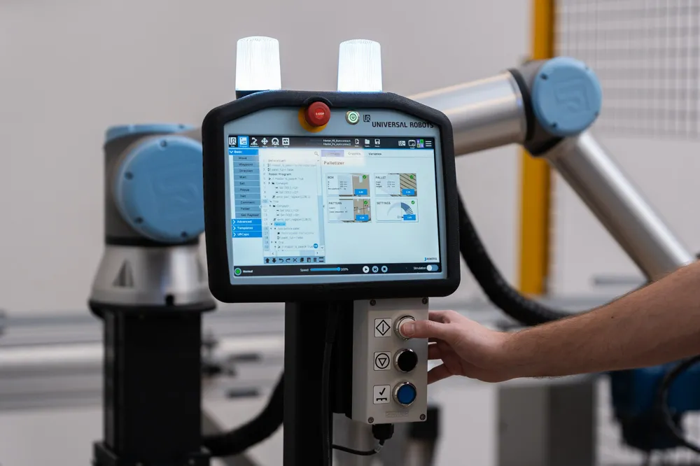 A Universal Robots teach pendant and a Robotiq operator button box on a palletizer pedestal, with the cobot arm behind.