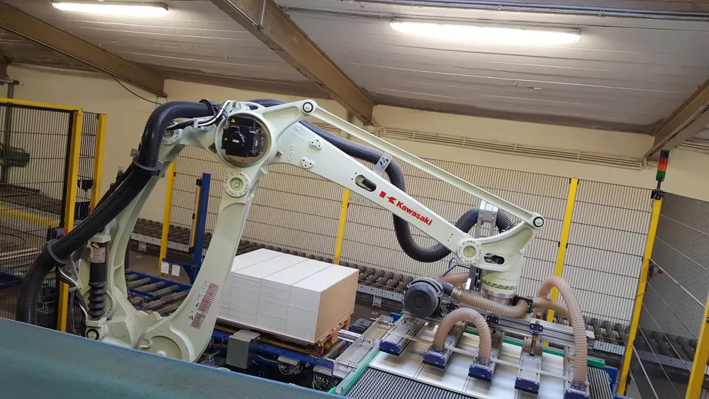 A white Kawasaki palletizing robot with a vacuum gripper stacking boards, inside yellow-posted mesh fence panels with a signal light