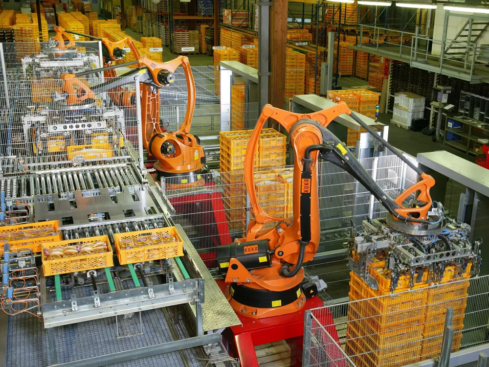Orange KUKA robots stacking yellow bread crates onto pallets inside mesh fencing at a bakery.