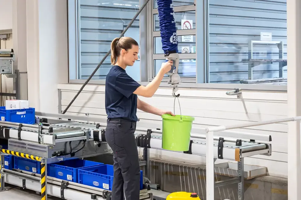 A worker lifting a green bucket with an overhead blue vacuum tube lifter next to a roller conveyor.
