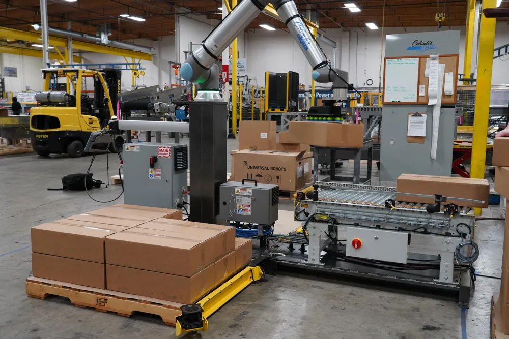 A cobot arm on a column next to a roller conveyor, stacking cardboard cases on a wooden pallet in a factory.