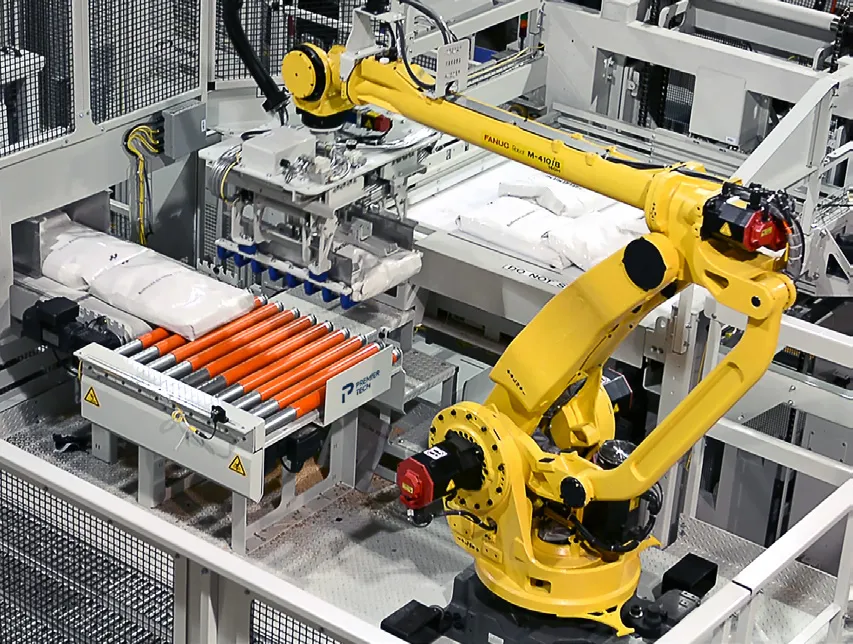 A yellow robot arm placing white bags onto a roller table inside a guarded palletizer.