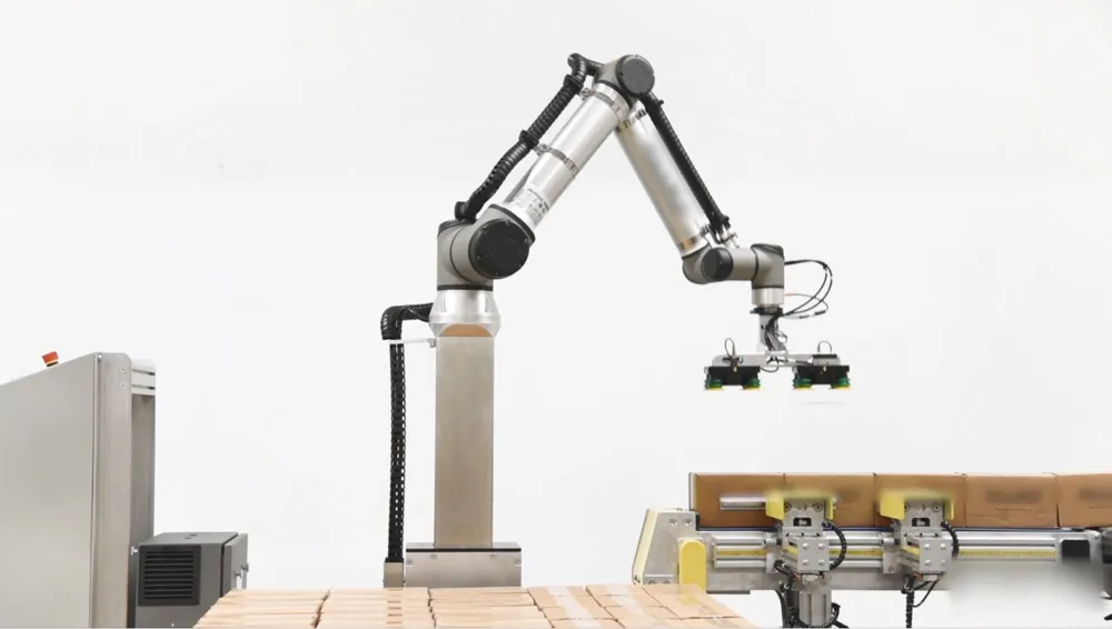 A Universal Robots cobot arm on a tall column with a two-zone vacuum gripper over a layer of boxes, next to an infeed conveyor.