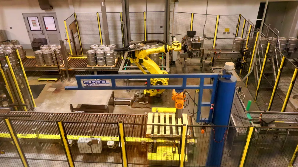 A yellow robot arm inside a fenced cell moving beer kegs from a conveyor to pallets.