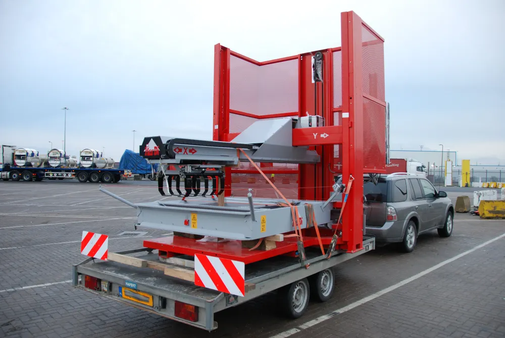 A red layer palletizer strapped to a flatbed trailer behind a car in a port parking lot.